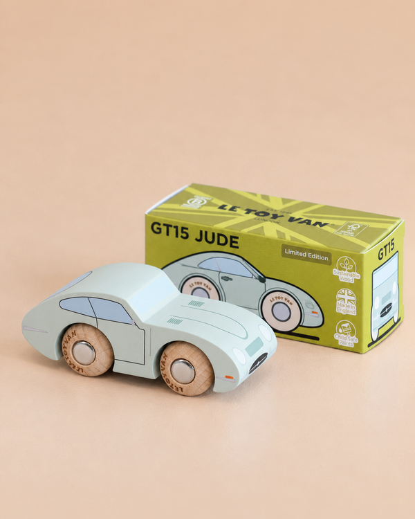 A light blue vintage toy car with wooden wheels sits in front of its green and yellow box labeled “No. 2 GT15 Jude | Made in England.” The background is a plain, soft beige surface, making this charming wooden car an ideal gift for kids.
