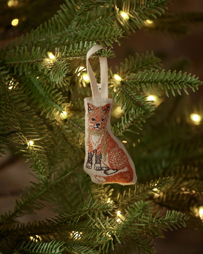 The Coral & Tusk Fox Tree Trimmer Ornament showcases an embroidered fox hanging from a green tree branch with warm white lights—ideal as a festive holiday gift or a cute addition to your tree décor.
