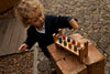 A young child with curly hair plays outdoors on a stone and wood surface with the Rainbow Pound -a- Peg, a colorful wooden Montessori toy featuring vibrant pegs on a wooden stand.