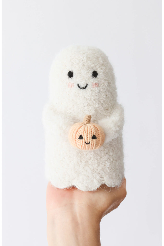 A hand holds a Cuddle + Kind Baby Ghost with Pumpkin, featuring a smiling face, rosy cheeks, and clutching a knitted pumpkin. Made from soft Peruvian cotton yarn. Plain white background.