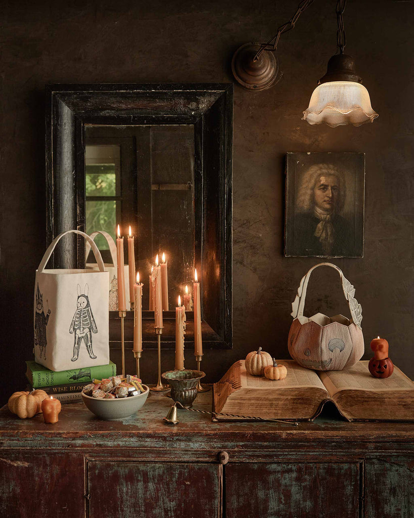 A rustic table holds a bowl of snacks, decorative pumpkins, the Coral & Tusk Jack-o'-Lantern Basket (ships in one week), and a basket. Lit candles sit on stacked books before a mirror. A vintage portrait hangs on a dark wall under a hanging lamp.