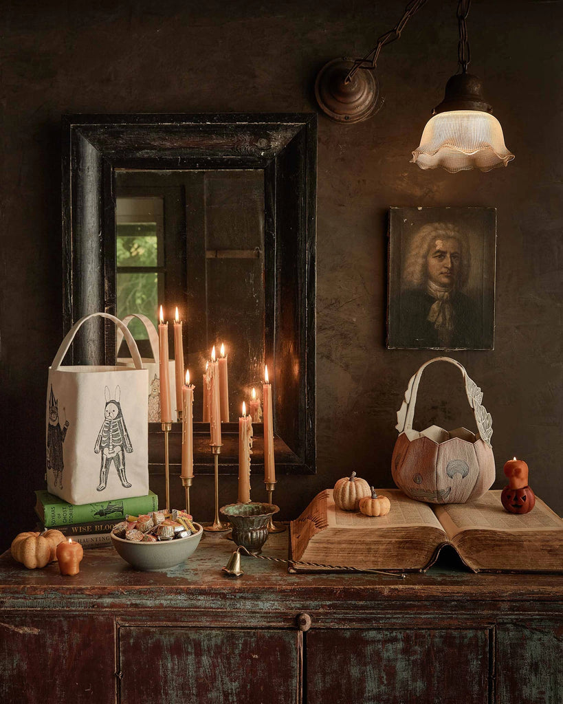 A rustic table with mini pumpkins, snacks, candles, the Coral & Tusk Halloween Trick Or Treat Bag (ships in one week), and a pumpkin basket sits before a dark wall with a mirror, old book, and portrait under a vintage lamp.