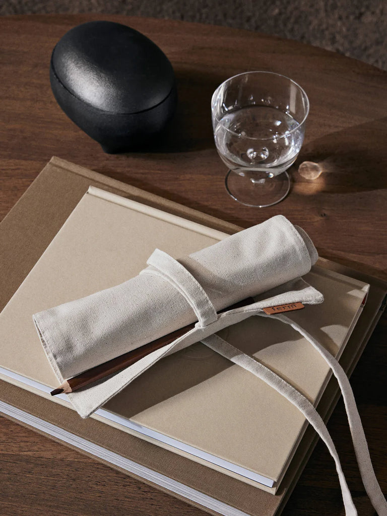 A glass of water, a black oval case, and stacked tan notebooks sit on a wooden table. On top of the notebooks is a Ferm Living Ally Pencil Wrap in off-white with a pencil partially tucked inside.