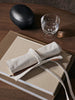 A glass of water, a black oval case, and stacked tan notebooks sit on a wooden table. On top of the notebooks is a Ferm Living Ally Pencil Wrap in off-white with a pencil partially tucked inside.
