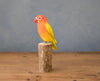 The Hand Carved Ornament - Lovebird - 7.5"H features a vibrant yellow and orange bird, handcrafted from sustainable softwood, perched on a wooden stump against a simple background on a wooden surface.