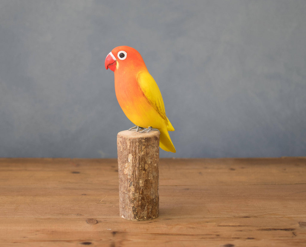 The Hand Carved Ornament - Lovebird - 7.5"H features a vibrant yellow and orange bird, handcrafted from sustainable softwood, perched on a wooden stump against a simple background on a wooden surface.