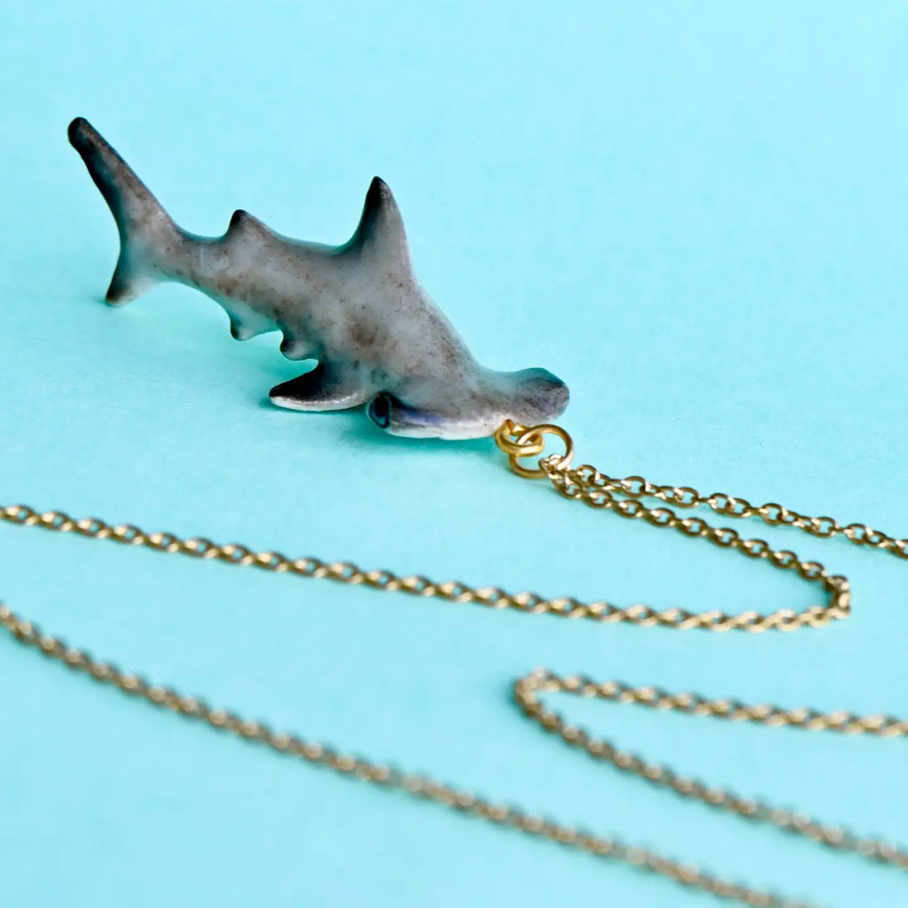 A close-up image of a Hammerhead Shark Necklace on a gold chain set against a light blue background. The pendant, resembling a hammerhead shark, has a realistic texture and detail, crafted from high quality porcelain. The delicate gold chain is partially coiled, adding elegance to these whimsical wearables.