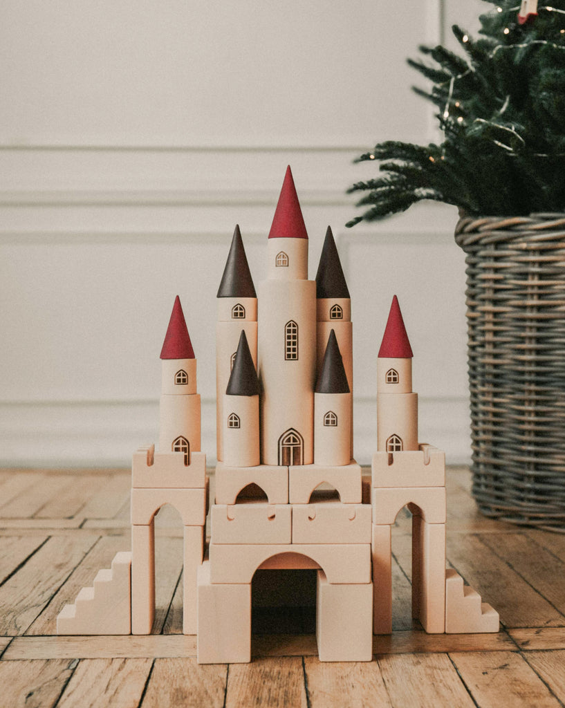 The Great Kingdom Building Set, a charming wooden toy castle with red and brown conical roofs, rests on a wooden floor. The pieces are designed with smooth edges and are coated with non-toxic paint. In the background, a wicker basket holds a green plant gently adorned with twinkling lights.