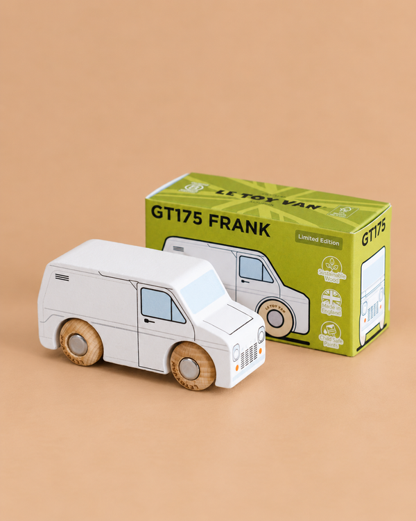 A classic toy van, crafted as a wooden white van with charming wooden wheels, sits in front of a green box labeled “No. 32 GT175 Frank | Made in England.” The van illustration on the box matches the toy’s design, inviting imaginative play. The background is beige.