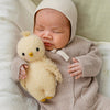 A baby in a beige knit outfit and bonnet lies on a light blanket, holding a soft yellow Cuddle + Kind Baby Bouclé Chick (Buttercup) collectible stuffed animal.