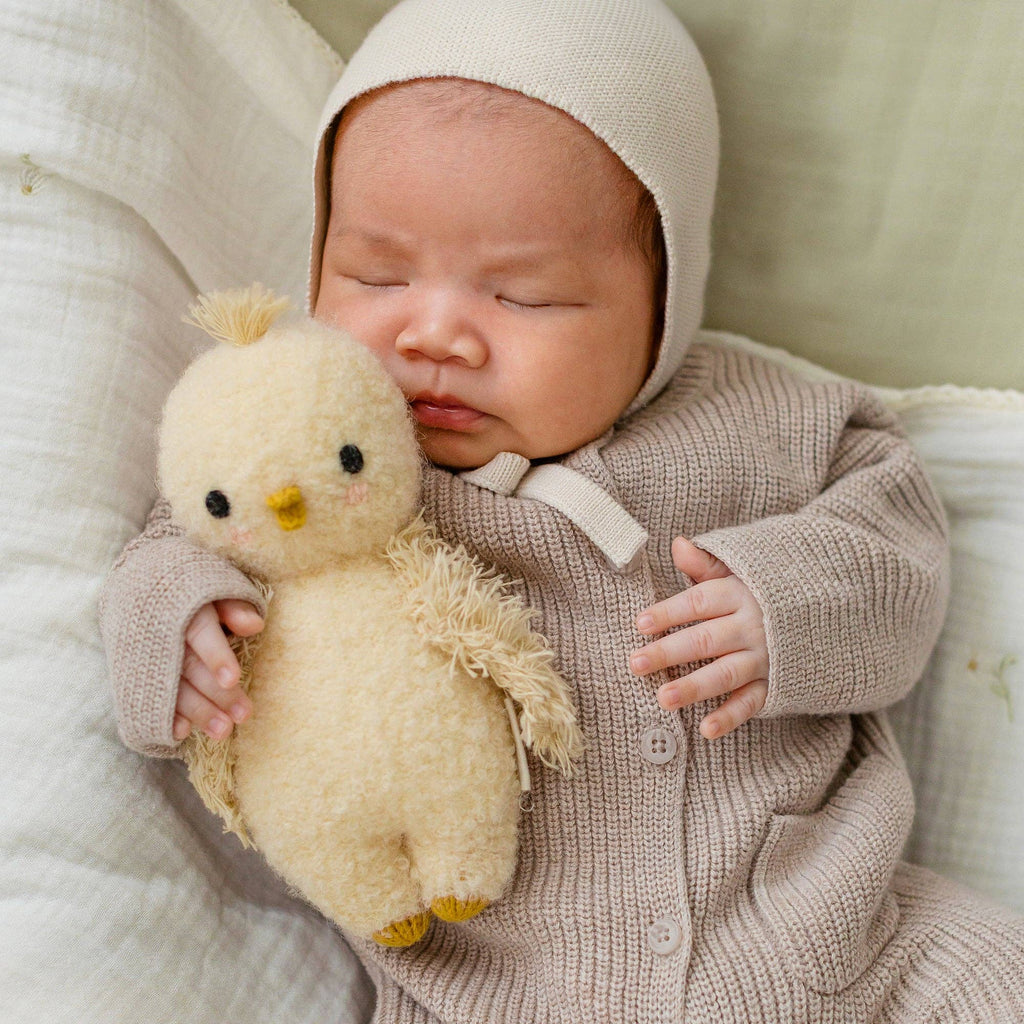 A baby in a beige knit outfit and bonnet lies on a light blanket, holding a soft yellow Cuddle + Kind Baby Bouclé Chick (Buttercup) collectible stuffed animal.