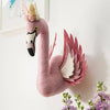 The Handmade Felt Flamingo with Wings is a plush pink wall decor, featuring felt wings and a flower crown. Mounted on a white wall near light purple flowers, it adds charm alongside framed artwork in the background.