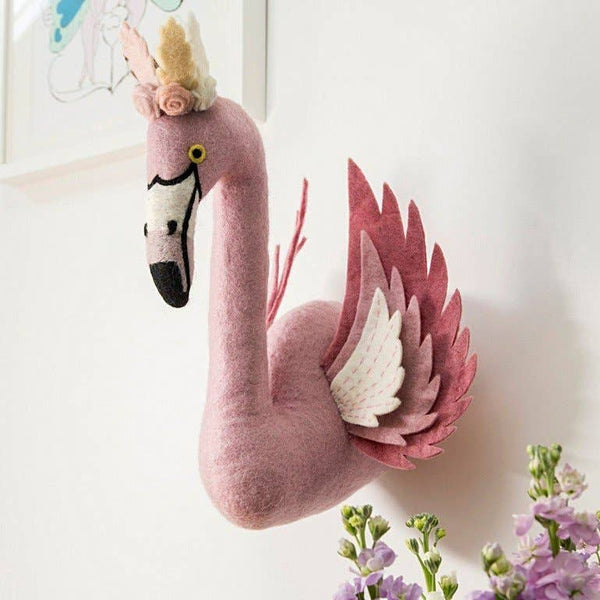 The Handmade Felt Flamingo with Wings is a plush pink wall decor, featuring felt wings and a flower crown. Mounted on a white wall near light purple flowers, it adds charm alongside framed artwork in the background.