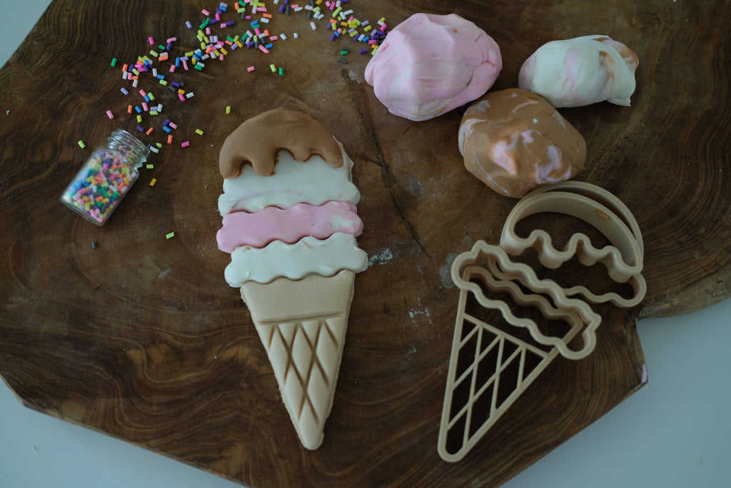 Experience hours of sensory play with the Ice Cream Cutter Set - Eco Playdough Tool, featuring a wooden ice cream cone-shaped sugar cookie design topped with frosting, colorful sprinkles, and playful marbled dough balls. Includes an eco-friendly ice cream playdough cutter and a small bottle of sprinkles—perfect for creative activity on any wooden surface.