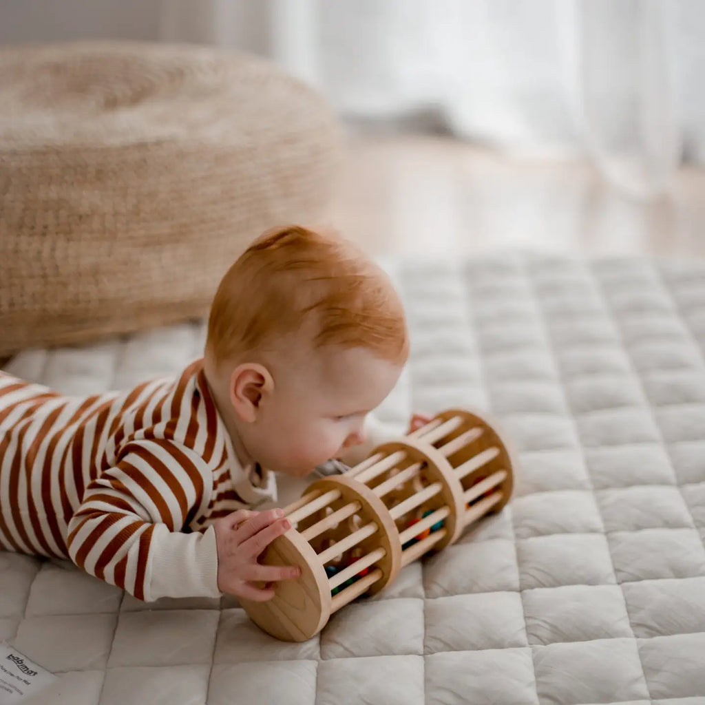 A baby with red hair, wearing a striped onesie, is lying on a quilted mat and playing with a non-toxic Wooden Rain Maker. The background features a woven basket and a soft, light-colored ambiance.