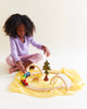 A young girl with curly hair, in a light purple top and pink pants, sits on a Sarah's Silks Solid Color Playsilk - Sunshine, enjoying open-ended play with a wooden train set, curved tracks, arches, and a toy tree.
