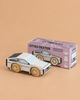 A sleek white wooden toy car with black detailing, the No. 23 GT130 Dexter | Made in England, sits in front of a purple box on a beige surface. The box displays an illustration matching this modern collectible and is labeled “GT130 DEXTER Limited Edition,” inviting young drivers to embark on imaginative adventures with this futuristic wooden car.