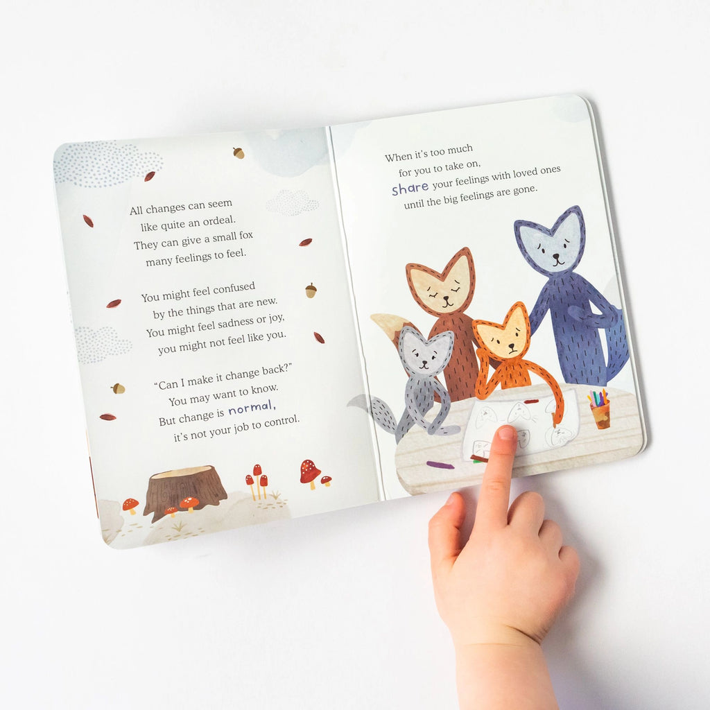 A child's hand pointing to a page in the Slumberkins Fox Kin + Lesson Book On Family Change featuring illustrated cats with a backdrop of trees. The open book, made with hypoallergenic fiberfill, lies on a white surface.
