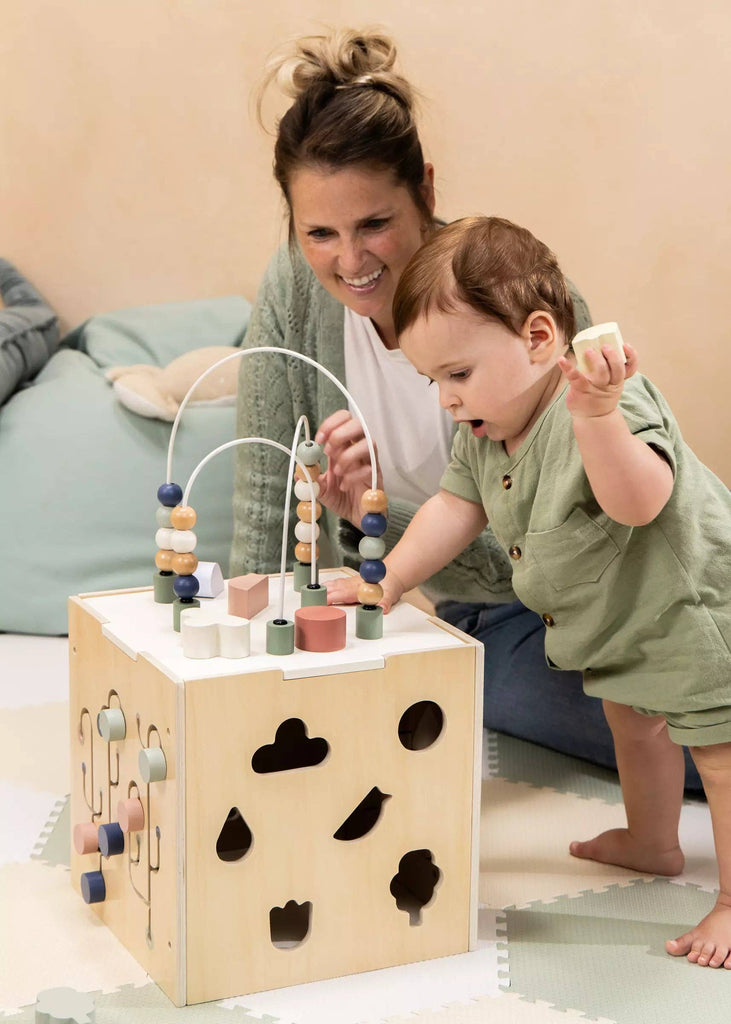 A woman and a toddler play with the Wooden Activity Cube, featuring bead mazes and shape sorters, on a soft mat in a cozy room—encouraging fine motor skills development.