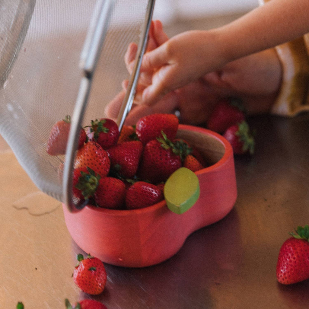 Someone pours freshly washed strawberries from a metal strainer into the Pepa the Apple Bowl & Spoon Set, which is pink and green, while some strawberries are also on the table.