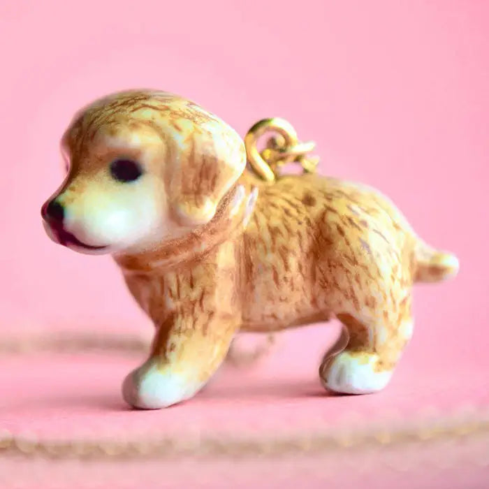 A small, fine porcelain figurine of a Golden Retriever Necklace with a pink background. The puppy has a shiny texture and a loop on its back, possibly for hanging as a decoration.