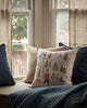 A cozy window seat with a blue quilt, blue and beige pillows, and the Coral & Tusk Winter Fun Pillow, featuring an embroidered winter scene. Soft daylight filters through beige curtains and large windows—a festive touch for holiday decor.