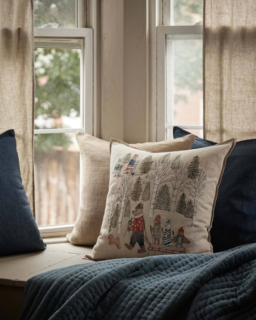 A cozy window seat with a blue quilt, blue and beige pillows, and the Coral & Tusk Winter Fun Pillow, featuring an embroidered winter scene. Soft daylight filters through beige curtains and large windows—a festive touch for holiday decor.
