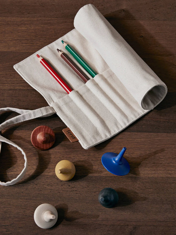 The Ferm Living Ally Pencil Wrap in off-white, filled with red, green, and brown pencils, sits on a wooden table beside four colorful spinning tops in blue, yellow, white, and brown.