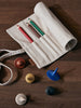 The Ferm Living Ally Pencil Wrap in off-white, filled with red, green, and brown pencils, sits on a wooden table beside four colorful spinning tops in blue, yellow, white, and brown.