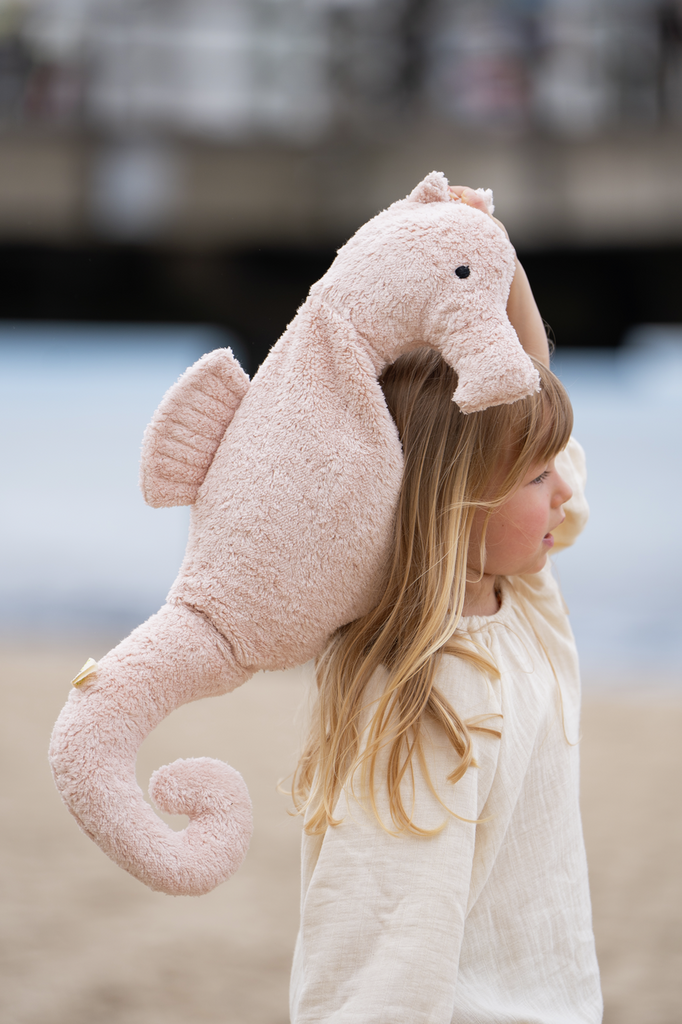 A young child with long blonde hair stands on a sandy beach, holding a large Senger Naturwelt Cuddly Animal - Seahorse plush toy over their shoulder.