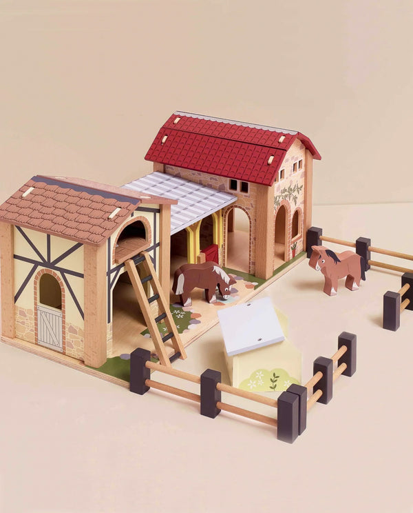 Wooden Farmyard Stables includes two stables, a fenced area, ladder, two toy horses, and a feeding station. Features red and brown barn roofs with painted details—perfect for imaginative small world play.