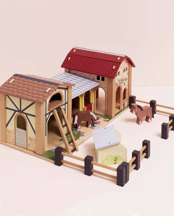 The Wooden Farmyard Stables includes two barn buildings, a fenced area, two toy horses, ladders, and a small trough—all made from FSC-certified wood and displayed on a light surface.