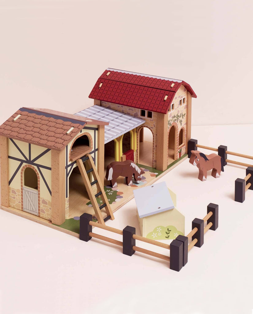 The Wooden Farmyard Stables includes two barn buildings, a fenced area, two toy horses, ladders, and a small trough—all made from FSC-certified wood and displayed on a light surface.