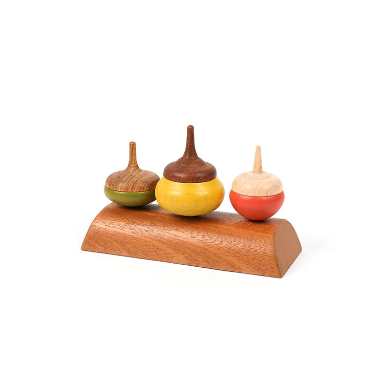 The Wooden Spinning Top Set - Chestnut features three colorful tops in green, yellow, and red, made from sustainable wood and displayed upright on a smooth, curved wooden stand against a white background.