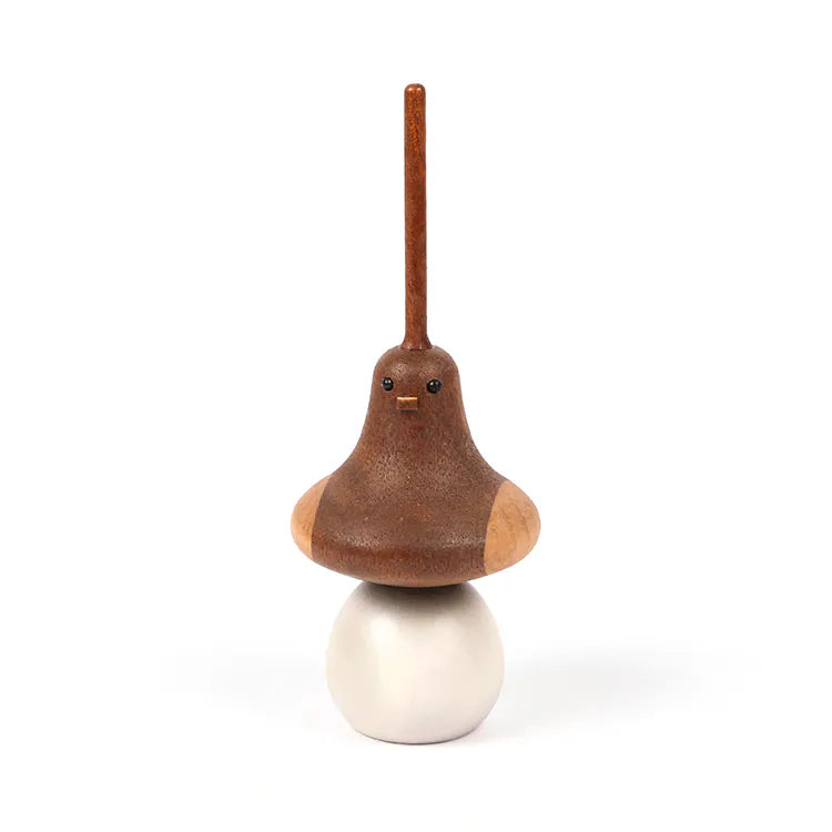 The Wooden Spinning Top - Bird features a long, upright stick as its head, small round eyes, and wing-like sides. Crafted from sustainably sourced wood, it sits perched on a smooth white sphere against a plain white background.