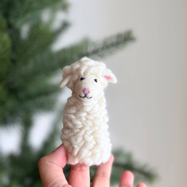 A hand holds the Felt Finger Puppet: Fluffy Sheep - White, featuring a pink nose, black eyes, and a small smile, in front of a blurred green pine tree—perfect for imaginative play or storytelling.