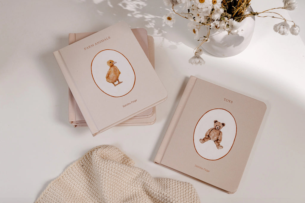 Two illustrated board books from the Our Little Library Book Box Set titled "farm animals" and "toys" on a white surface, with a dried flower arrangement and a beige knitted fabric next to them. The books have drawings of a