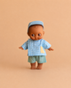 The Mini Pancha Doll - Noah with Blue Checkered Outfit features dark skin and comes dressed in a blue checkered shirt, green shorts, and a matching blue cap, standing barefoot against a solid beige background. Perfect for imaginative play and crafted from high-quality vinyl.