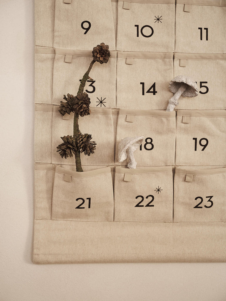 A Ferm Living Star Christmas Calendar - Sand/Black canvas calendar with 24 numbered pockets, decorated with a pinecone branch and two small mushroom ornaments.