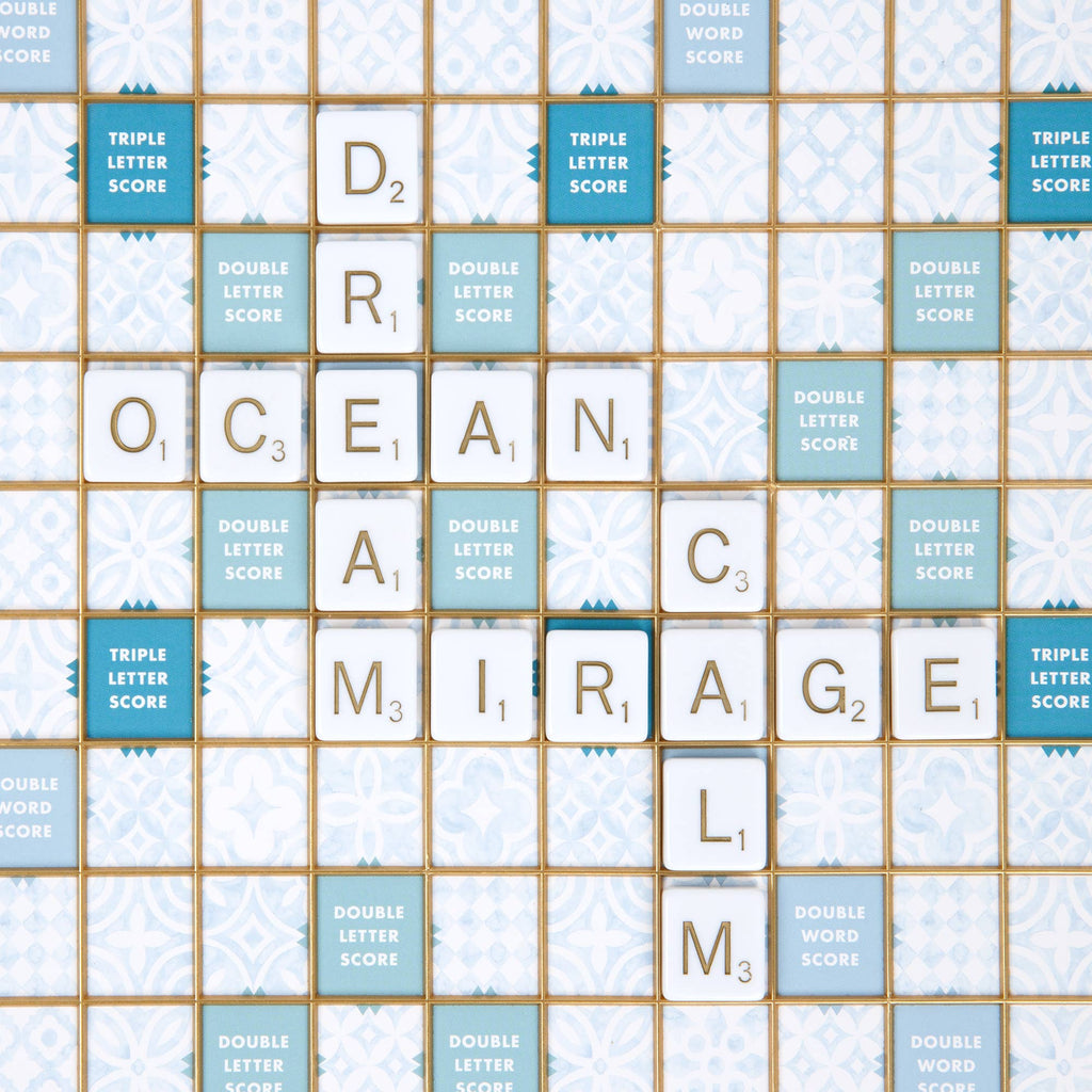 A Scrabble Bianco Edition game board featuring white tiles spelling “DREAM,” “OCEAN,” “MIRAGE,” and “CALM” arranged in a crossword pattern on a patterned blue and white grid.