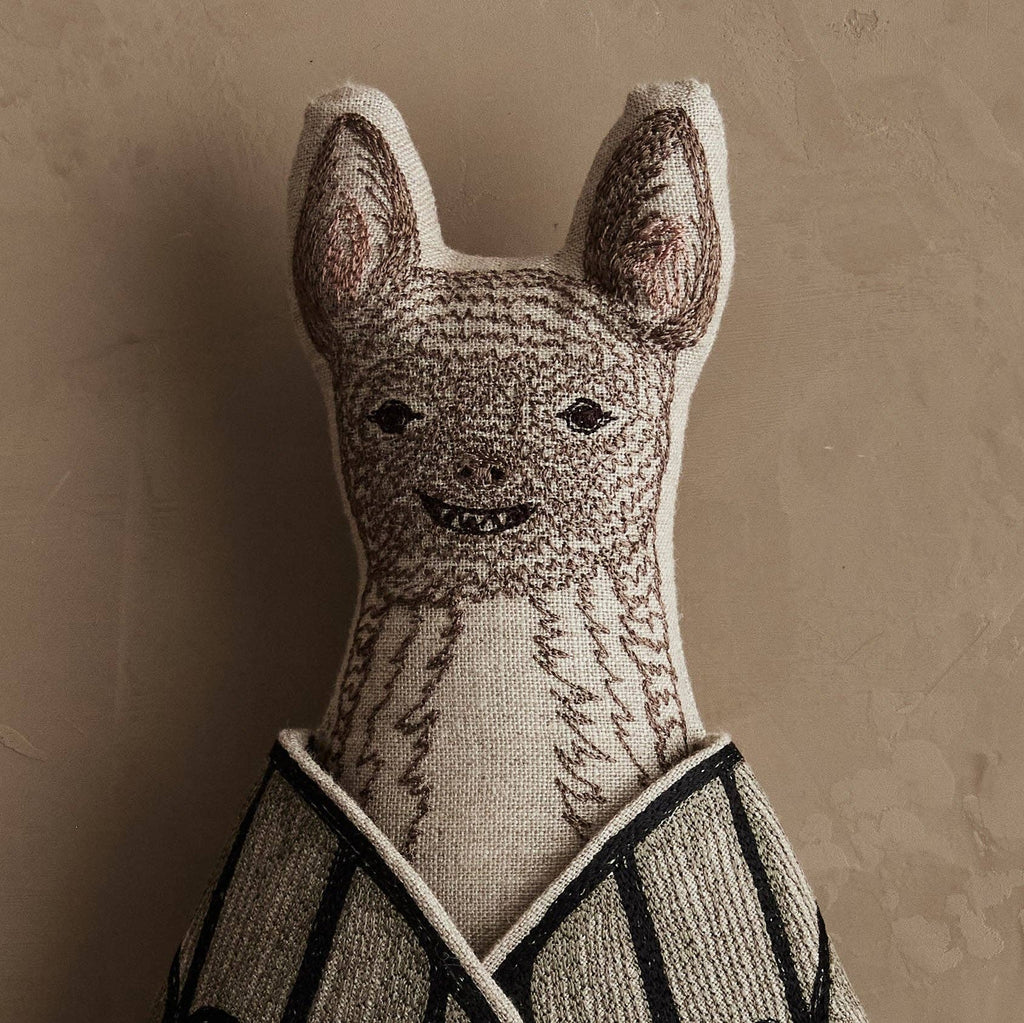 The Coral & Tusk Bat Surprise Doll is a decorative bat plush toy with embroidered facial features, pointy ears, fabric wings, and a striped cloak, set against a textured beige background. Ships in one week.