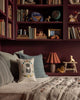 A cozy reading nook with maroon bookshelves filled with books and toys, a cushioned bench decorated with spring pillows—one featuring the Coral & Tusk Kitten Cameo Pillow—a taupe quilt, a small lamp, and wooden animal figurines.