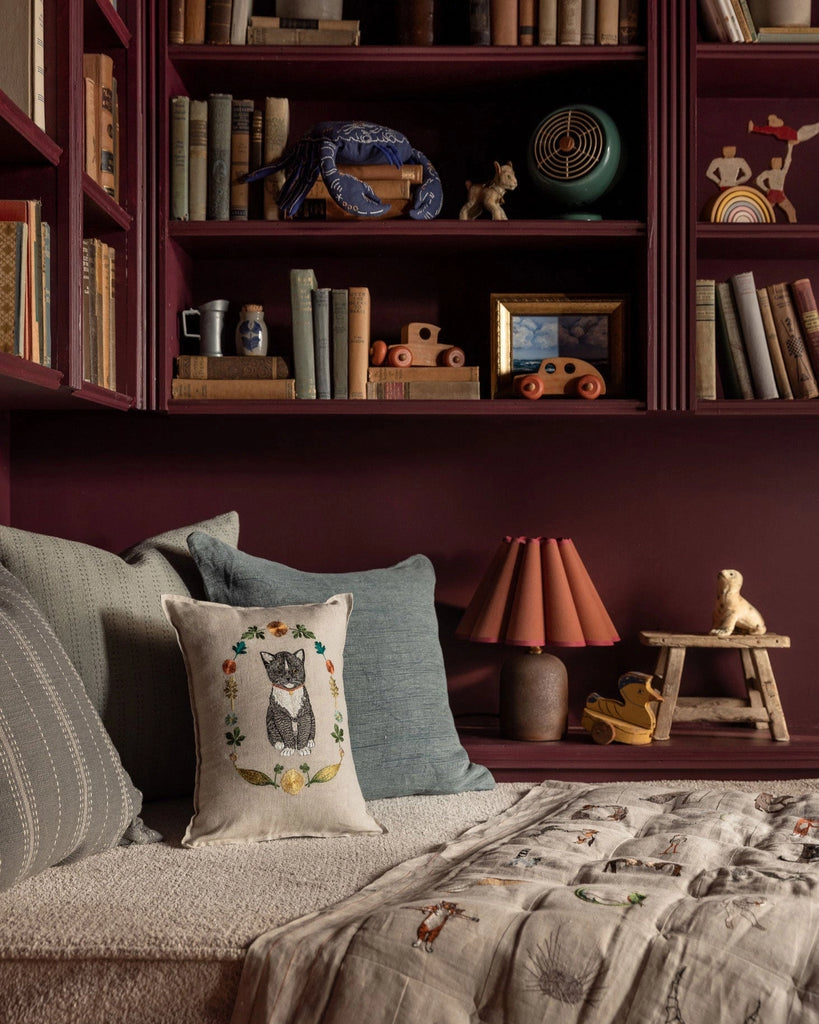 A cozy reading nook with maroon bookshelves filled with books and toys, a cushioned bench decorated with spring pillows—one featuring the Coral & Tusk Kitten Cameo Pillow—a taupe quilt, a small lamp, and wooden animal figurines.
