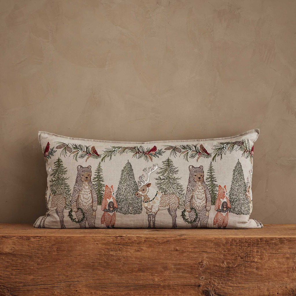 Coral & Tusk Christmas Cheer lumbar pillow featuring an embroidered winter woodland scene with bears, a reindeer, and foxes among pine trees and evergreen garlands.