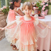 Two young girls, wearing matching Meri Meri Fairy Costumes with layered pink and cream designs and ruffled sleeves, are smiling and dancing in a bright room decorated with plants and a pink tablecloth. The girl in the front has her arms spread out, showcasing the flowy design of her fairy dress.