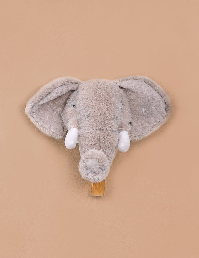 The Coat Hanger - Elephant features a plush gray elephant head with big floppy ears and white tusks on a tan background, serving as both a cute soft toy wall decor and a functional coat hanger.