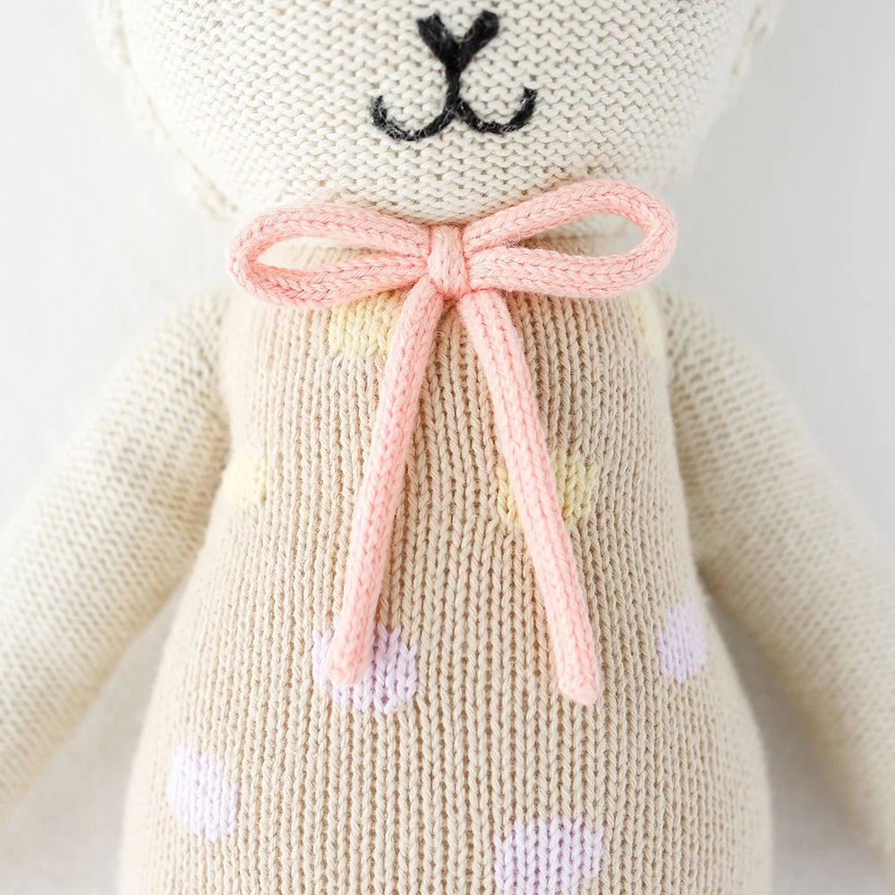 A close-up of Cuddle + Kind Lucy the Lamb, a fair trade hand-knit doll in cream-colored yarn with a pink bow and pastel polka dots, showing the lower face and upper torso.