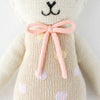 A close-up of Cuddle + Kind Lucy the Lamb, a fair trade hand-knit doll in cream-colored yarn with a pink bow and pastel polka dots, showing the lower face and upper torso.