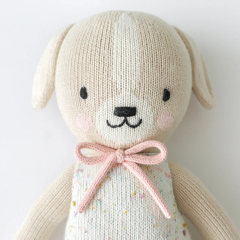 Cuddle + Kind Mia the Dog is a cream-colored, hand-knit stuffed doll made from premium cotton yarn, featuring black stitched eyes and nose, light pink cheeks, a pink bow, and a white speckled outfit.