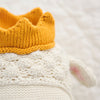 Close-up of a Cuddle + Kind Sebastian the Lamb knitted baby shoe with a yellow ribbed cuff, white textured body, and an ear-like detail. This sustainable, fair-trade piece appears against a soft, blurred background.